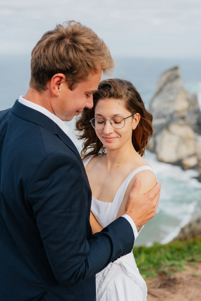 Detailní fotka usmívajících se novomanželů, kterým ve větru vlají vlasy na útesech u oceánu, Portugalsko.