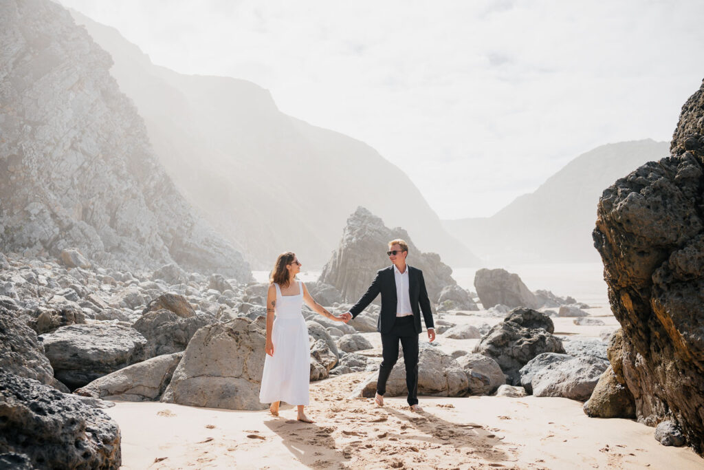 Uvolněné svatební focení na pláži: Ženich a nevěsta kráčí bosí v písku na Praia da Adraga.