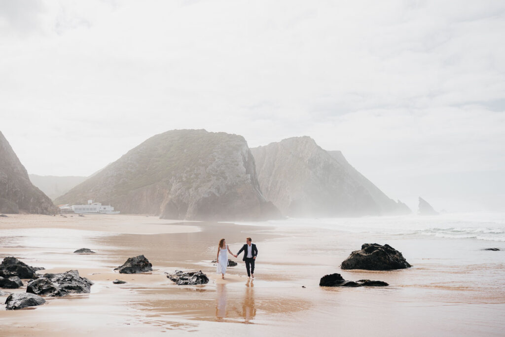 Romantická procházka po pláži ve svatebních šatech s vysokými skalami v pozadí.