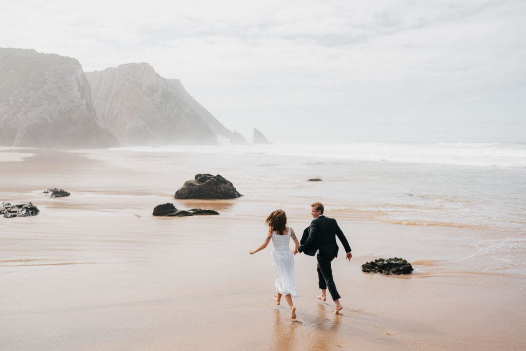 Ženich a nevěsta se drží za ruce a utíkají před vlnami na pláži v Portugalsku.